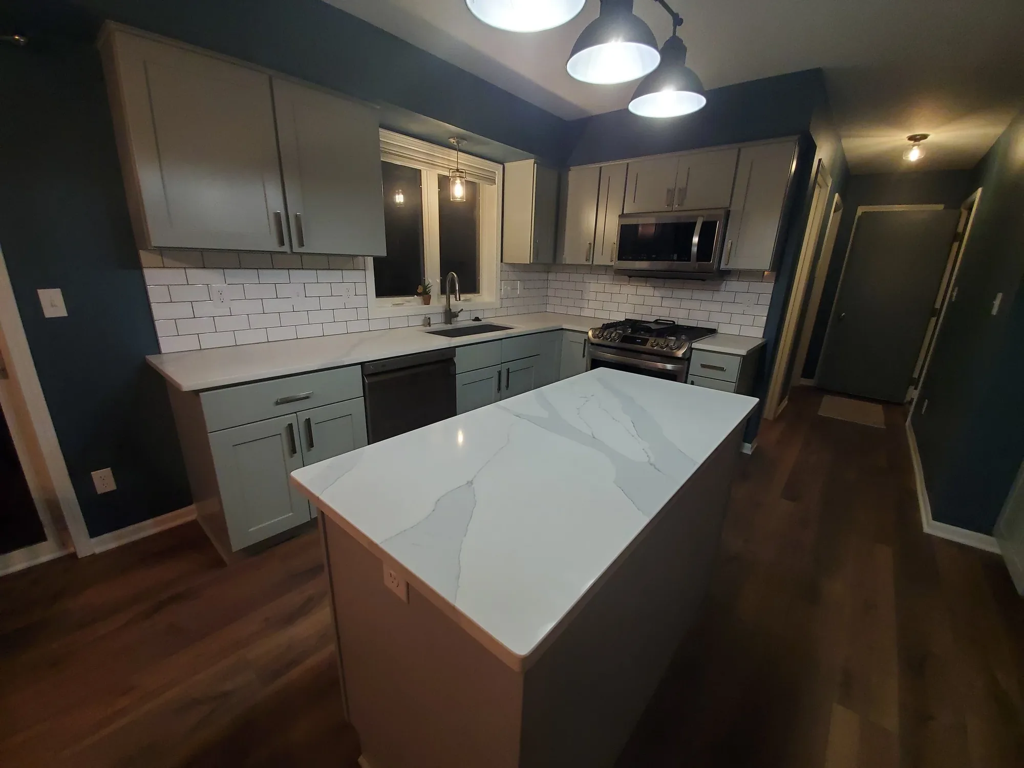 The same kitchen after remodeling, with grey cabinets and a quartz waterfall island